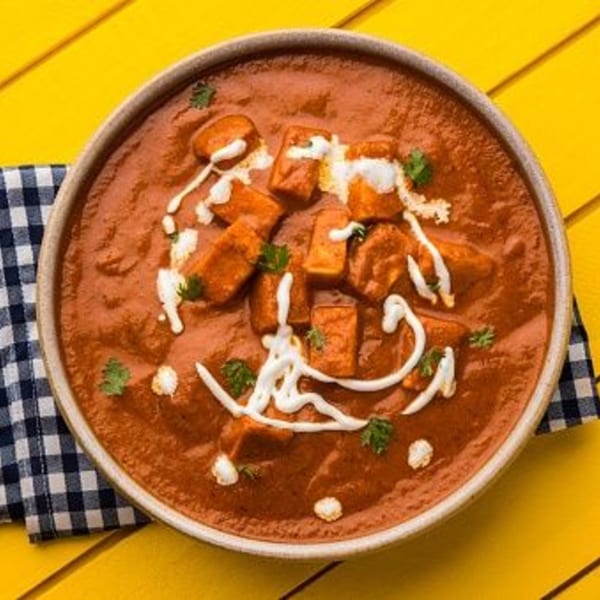 Paneer Butter Masala, a rich Indian curry with paneer cubes, cream, and cilantro, on a yellow table.