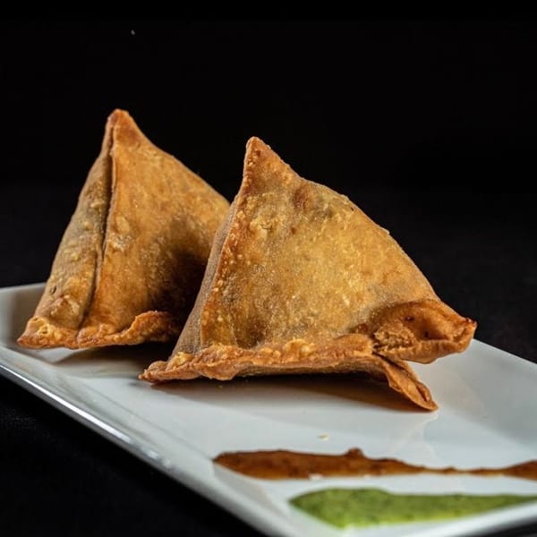 Two golden-brown samosas on a white plate with green and brown sauces, against a dark background.